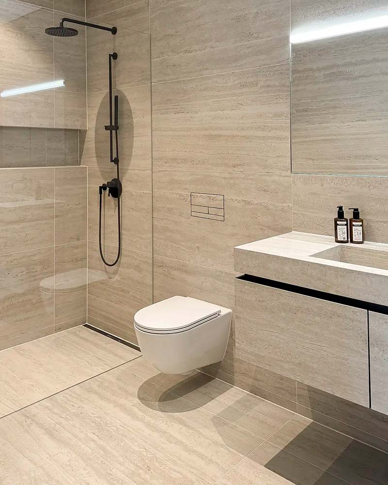 Modernes Badezimmer mit beiger Fliesenwand, wandhängendem WC, begehbarer Dusche mit schwarzer Armatur und Waschbecken mit zwei Seifenspendern.